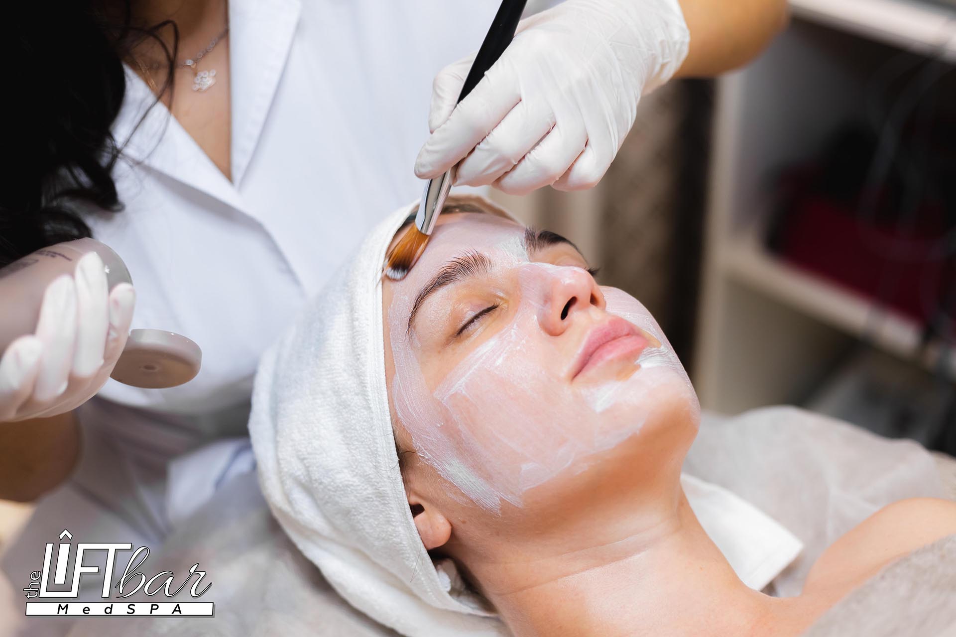 Beautician with a brush applies a white moisturizing mask to the face of a young girl client in a spa beauty salon anti aging treatment