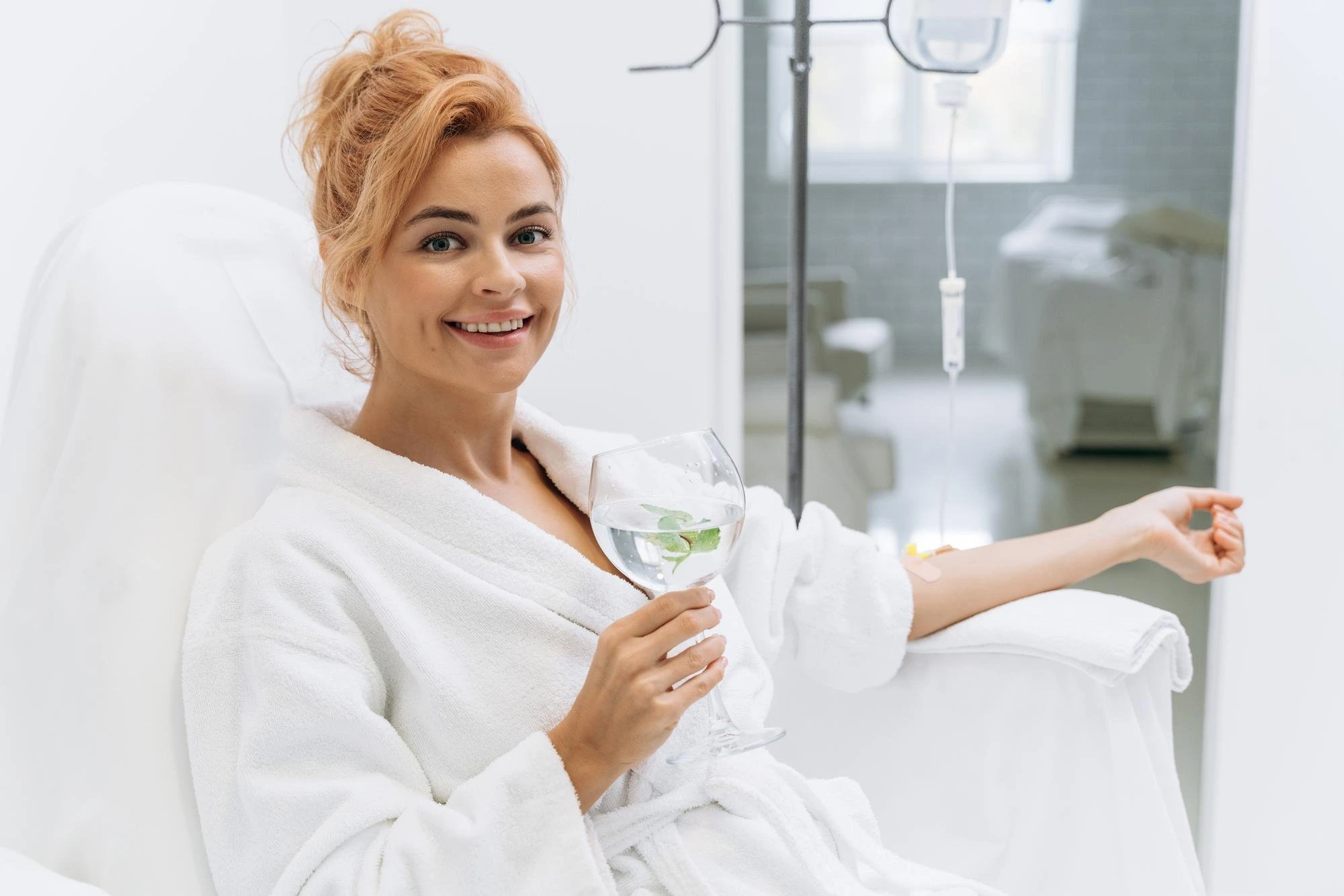 happy-woman-white-bathrobe-holding-glass-lemon-beverage-smiling-while-sitting-armchair-receiving-iv-infusion Benefits Of Myers Cocktail IV Therapy