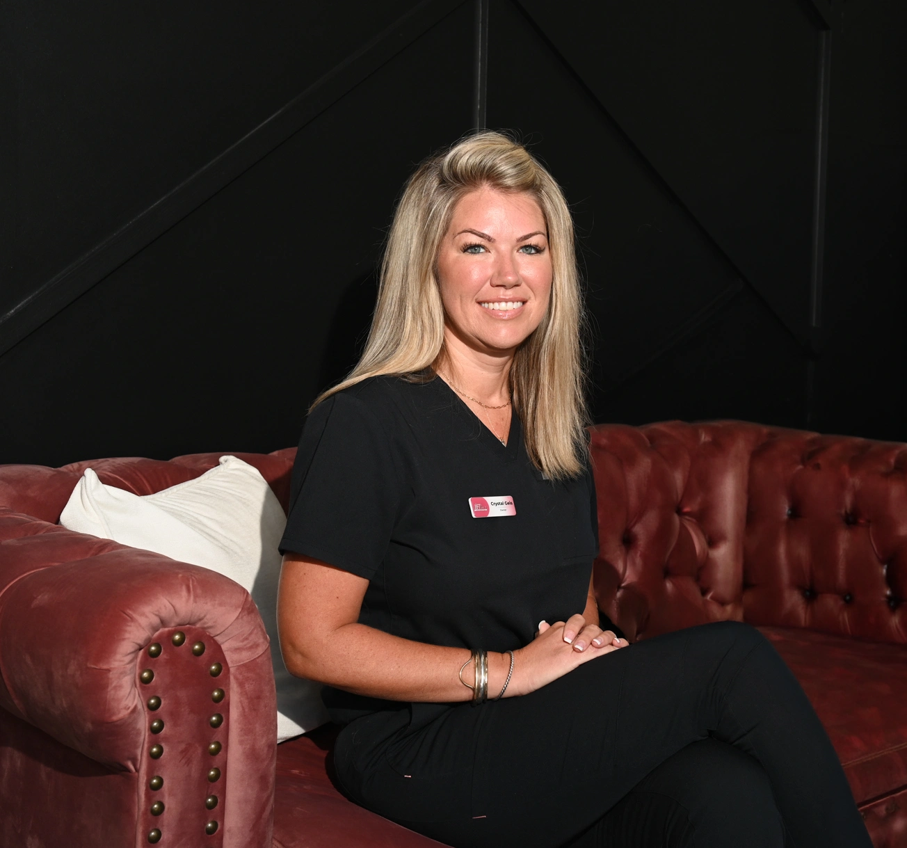 Smiling blonde woman in black scrubs sits on a red tufted sofa in a dark studio, wearing a name badge.